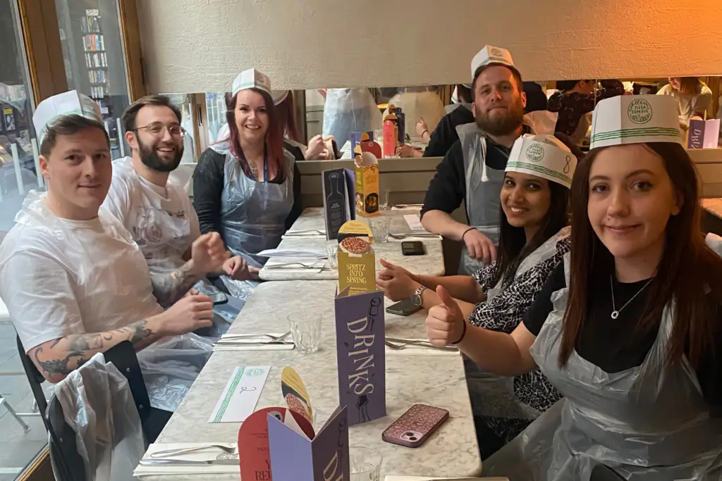 Group of people sat on long table wearing chef hats for pizza making social.