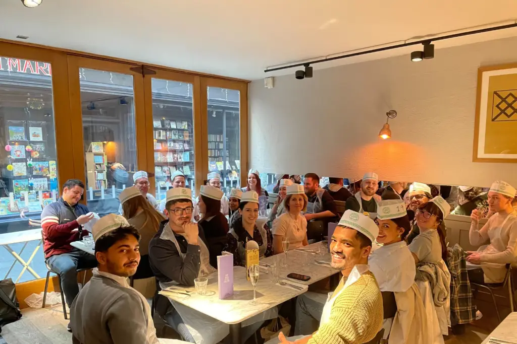 Large group of people sat across two tables in restaurant for pizza making social.