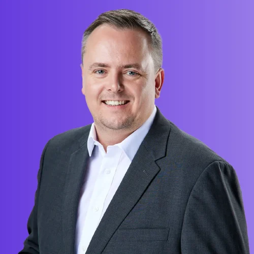 Image of Male, Chris Whitcombe, Managing Director of LDMS. Wearing white shirt and dark grey blazer, smiling into the camera against a purple gradient background.